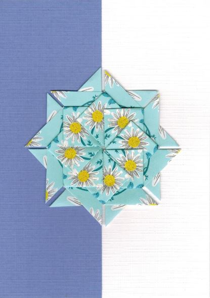 Een lichtblauwe rozet met witte bloemen op een kaart met bladwijzer motief.<br>Deze rozet is gemaakt uit Clipper calmer chameleon theezakjes.